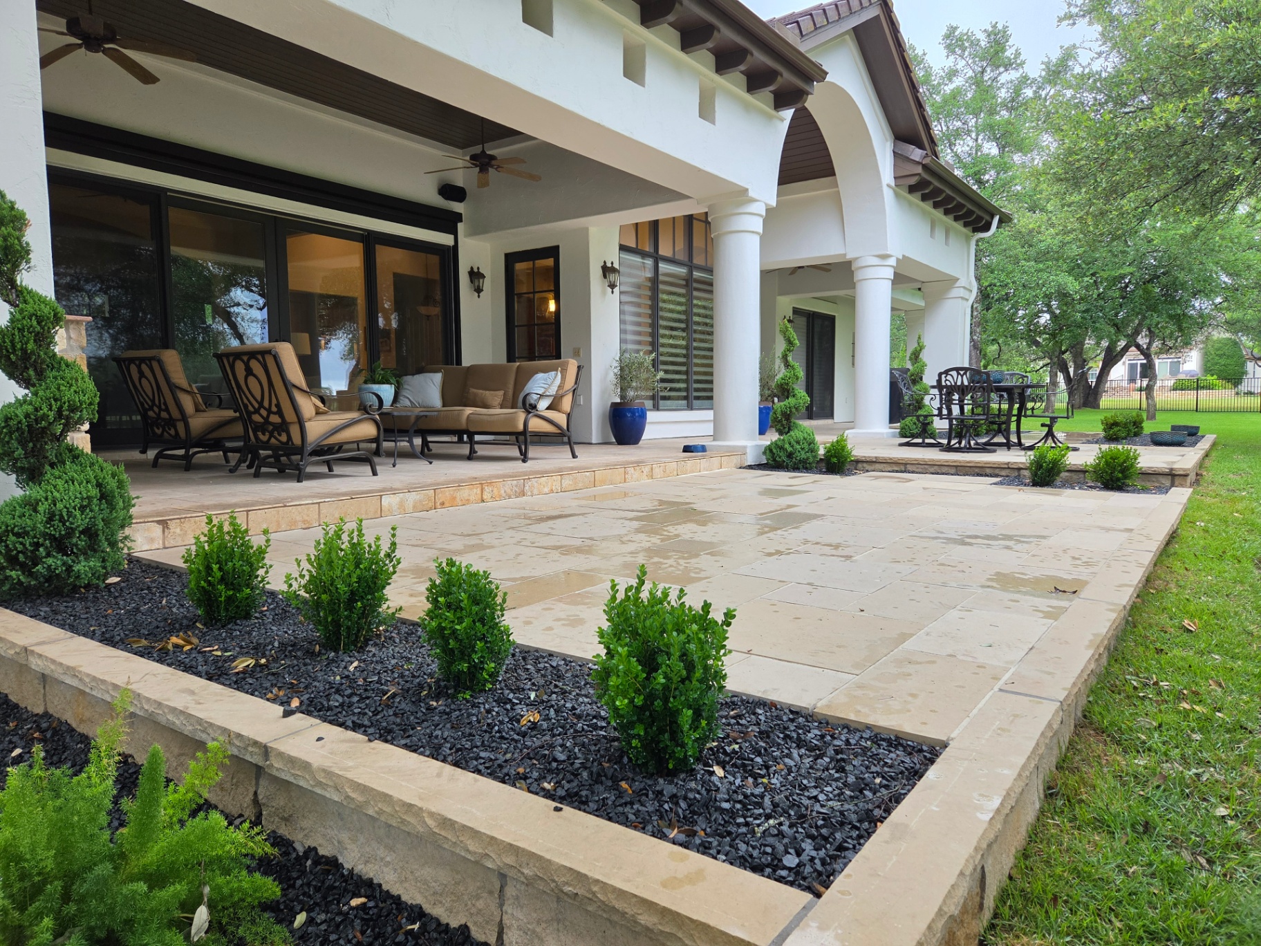 Covered patio with seating area, large stone patio, landscape beds, and mature trees